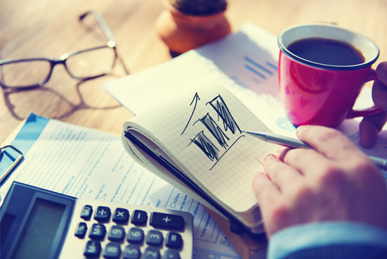 Coffee cup, notebook , paper with a chart on it, glasses, and calculator. Person's hand holds a pen.