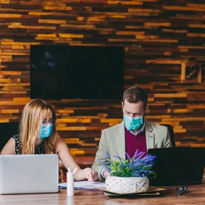 Two professionals sit around a table with their computers and face masks on, working together.