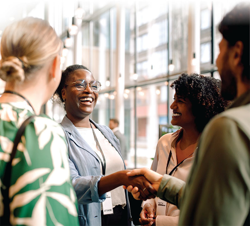 Four people standing and smiling in a bright, modern lobby. One woman is laughing and shaking hands with someone outside the frame, while the others look on, smiling.